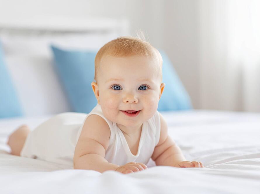 Μωρό ξαπλωμένο σε λευκό κρεβάτι με χαμόγελο, σε φωτεινό υπνοδωμάτιο.