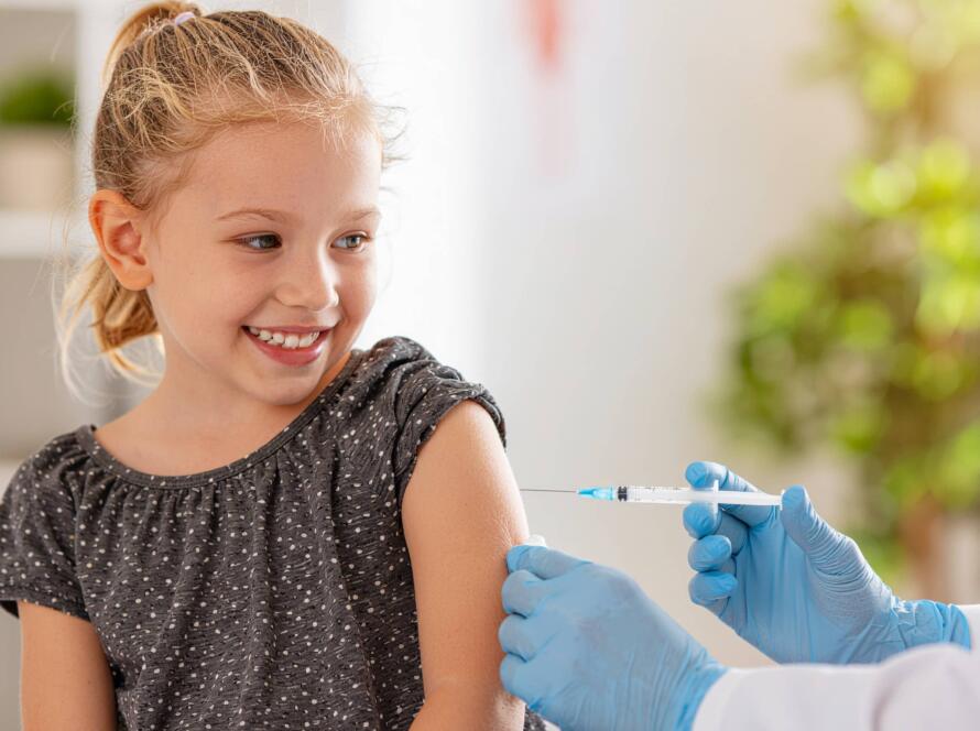 Κοριτσάκι χαμογελά καθώς δέχεται εμβόλιο στο χέρι από γιατρό με γάντια, σε ιατρικό περιβάλλον.