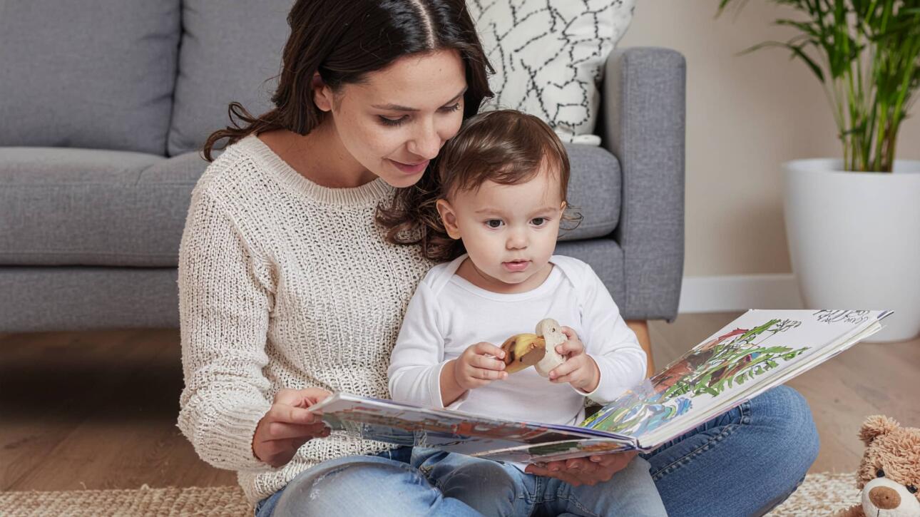 Μαμά διαβάζει βιβλίο σε μικρό παιδί καθισμένες στο πάτωμα του σαλονιού, με λούτρινα παιχνίδια δίπλα τους