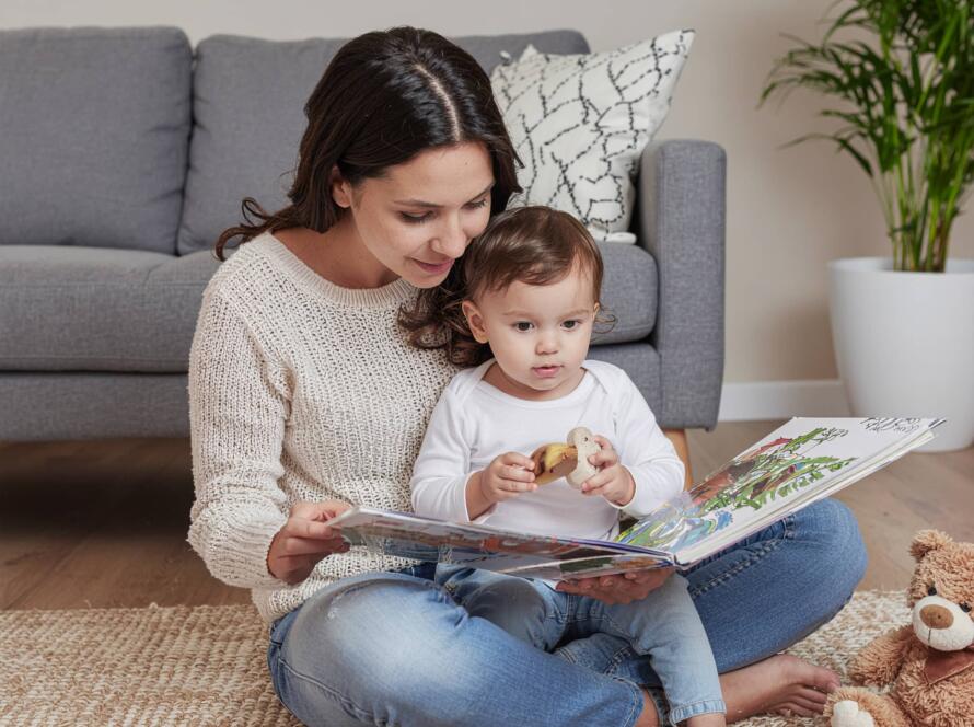Μαμά διαβάζει βιβλίο σε μικρό παιδί καθισμένες στο πάτωμα του σαλονιού, με λούτρινα παιχνίδια δίπλα τους