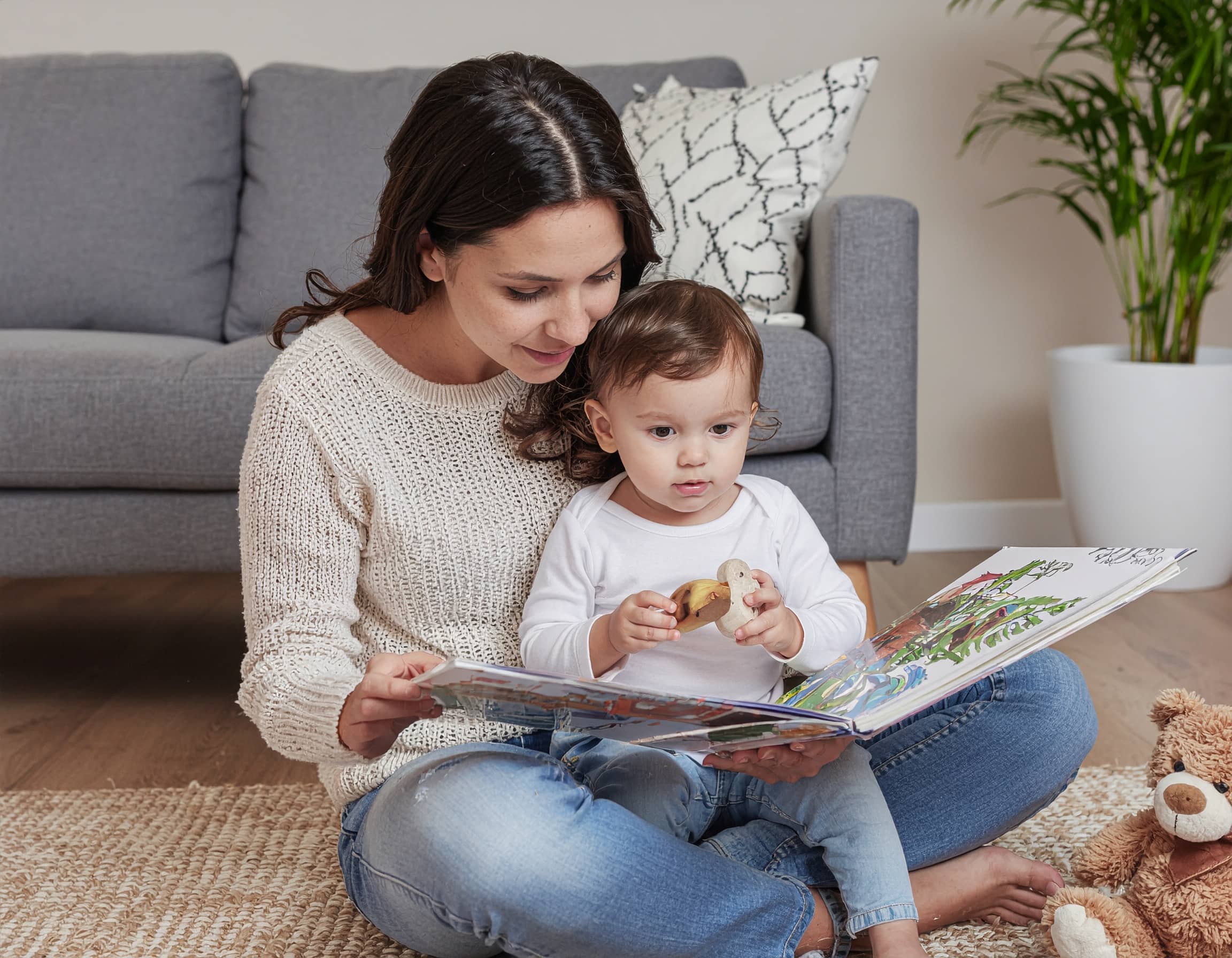 Μαμά διαβάζει βιβλίο σε μικρό παιδί καθισμένες στο πάτωμα του σαλονιού, με λούτρινα παιχνίδια δίπλα τους