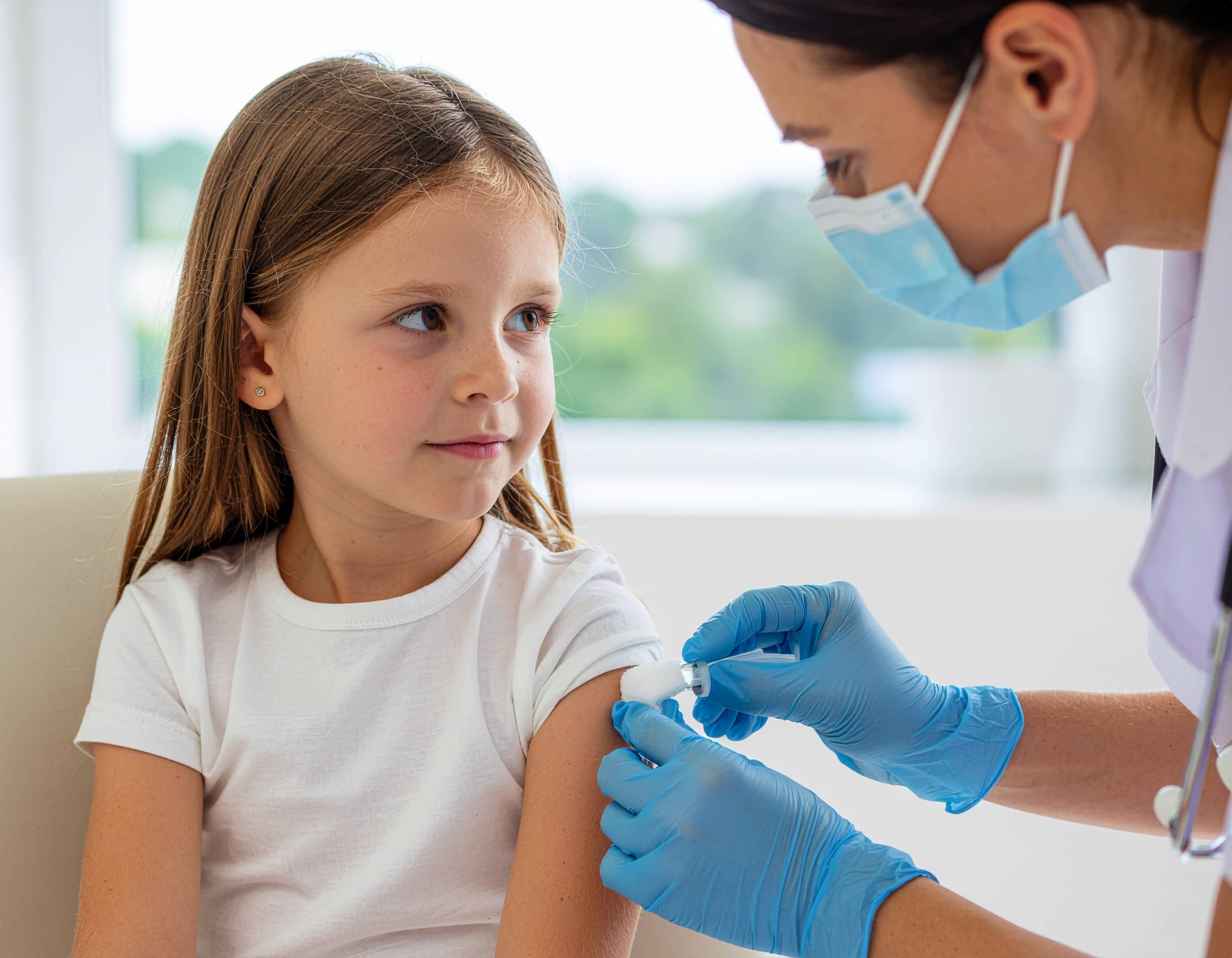 Νοσηλεύτρια με γάντια και μάσκα καθαρίζει το χέρι μικρού κοριτσιού πριν τον εμβολιασμό, σε ιατρικό περιβάλλον.