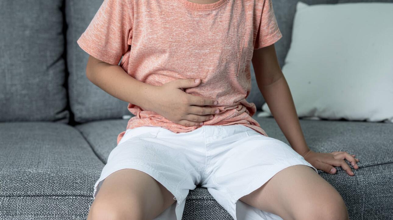 Αγόρι με πορτοκαλί μπλούζα και λευκό σορτς κάθεται σε καναπέ και κρατά την κοιλιά του, δείχνοντας ενόχληση ή πόνο.