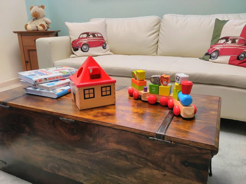 Ξύλινο τραπέζι με παιδικά εκπαιδευτικά παιχνίδια, βιβλία, ξύλινο σπιτάκι και πολύχρωμη ξύλινη αμαξοστοιχία, σε άνετο παιδικό χώρο αναμονής.​