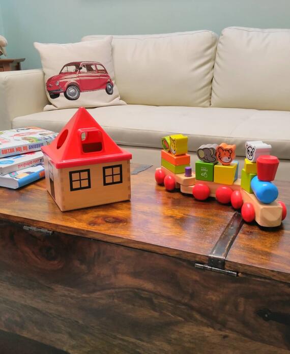 Ξύλινο τραπέζι με παιδικά εκπαιδευτικά παιχνίδια, βιβλία, ξύλινο σπιτάκι και πολύχρωμη ξύλινη αμαξοστοιχία, σε άνετο παιδικό χώρο αναμονής.​