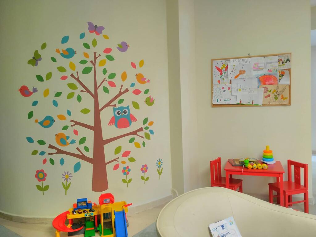 Τοίχος παιδικού χώρου διακοσμημένος με αυτοκόλλητο δέντρο και πουλάκια, παιδικό τραπεζάκι με κόκκινες καρέκλες και παιχνίδια, και πίνακας με ζωγραφιές παιδιών