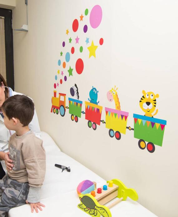 Παιδίατρος εξετάζει αγόρι σε εξεταστήριο διακοσμημένο με πολύχρωμο αυτοκόλλητο τρενάκι και παιχνίδια, δημιουργώντας φιλική ατμόσφαιρα