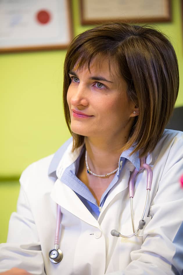 Η Παιδίατρος Αννα Βλάχου σε ιατρείο κάθεται και συνομιλεί με παιδί ή γονέα προβάλλοντας φροντίδα και επαγγελματισμό.